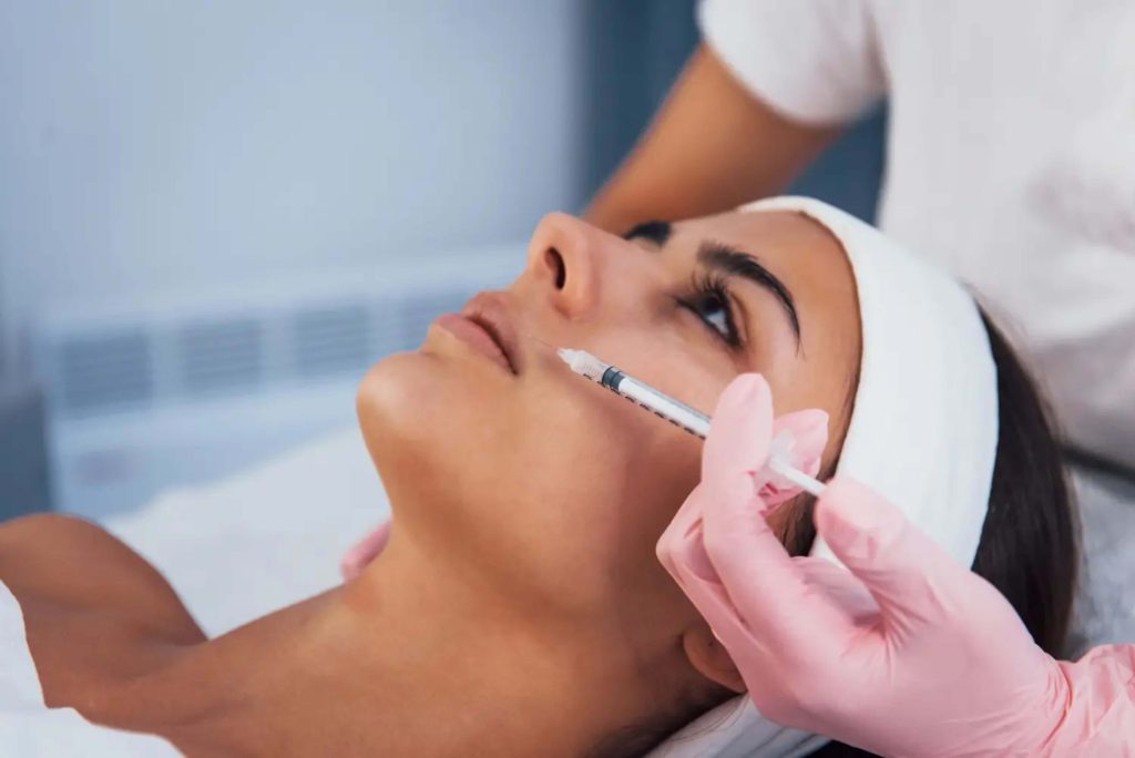 Medical professional administering facial injection treatment to female patient wearing white headband in clinical setting